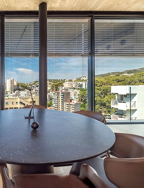 Round dark stone dining table surrounded by curved tan leather chairs in front of floor-to-ceiling windows overlooking the sea.