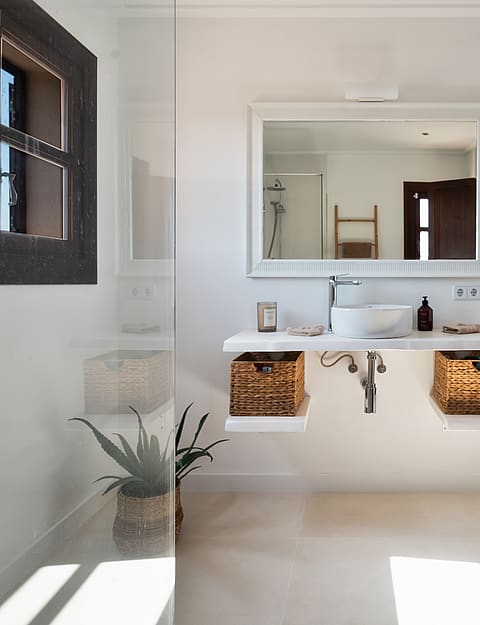Baño minimalista con mueble blanco suspendido, cestas de mimbre, espejo rectangular y ducha de cristal.