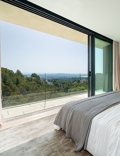 A modern master bedroom with a large bed, floor-to-ceiling sliding glass doors opening to a balcony with sea views.