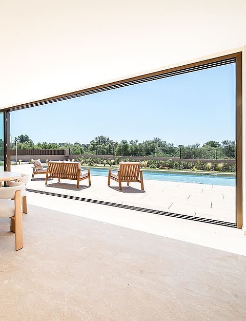 Dining area with oval light oak table and modern upholstered chairs opening onto a terrace with a swimming pool and trees.