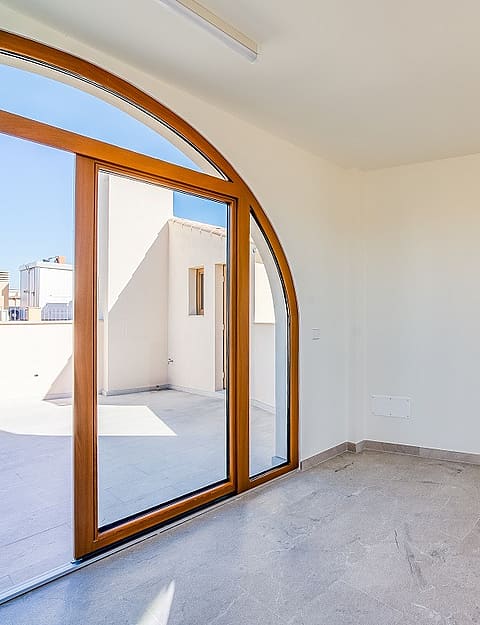A minimalist room with white walls, stone flooring, and a large wooden arched glass door leading to an outdoor terrace.