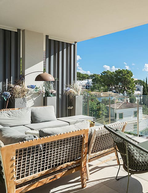 A covered terrace with grey outdoor furniture, glass railings, and a view of trees and houses under a clear blue sky.