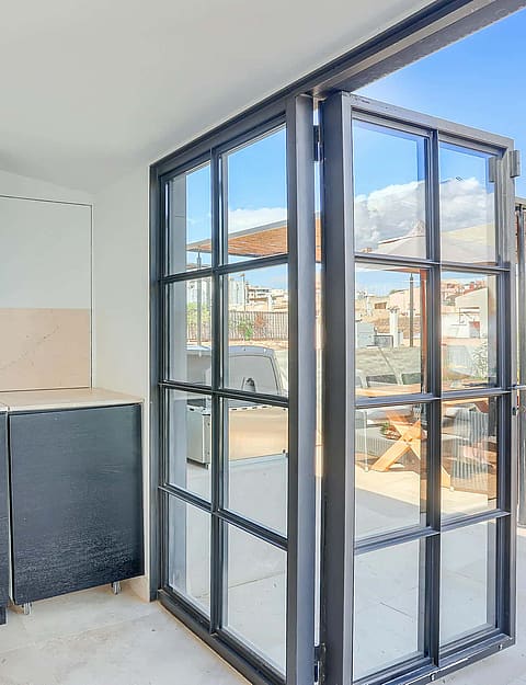 Compact outdoor kitchenette with charcoal cabinetry and stone backsplash adjacent to black-framed glass folding doors opening onto a terrace.
