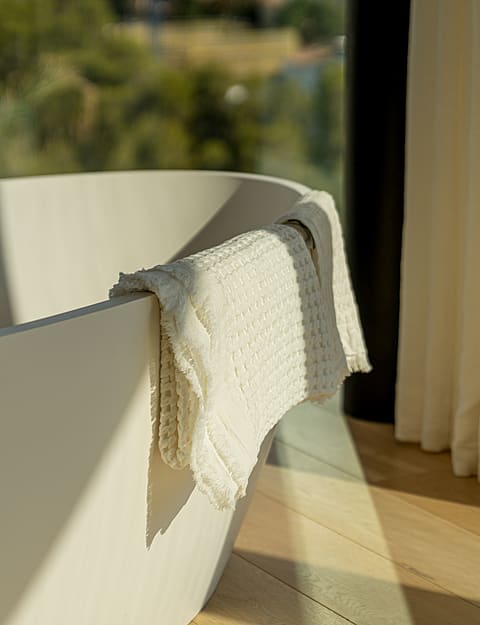 Close-up of a white freestanding bathtub with a textured waffle-weave towel draped over the rim, adjacent to floor-to-ceiling cream curtains and light oak flooring.