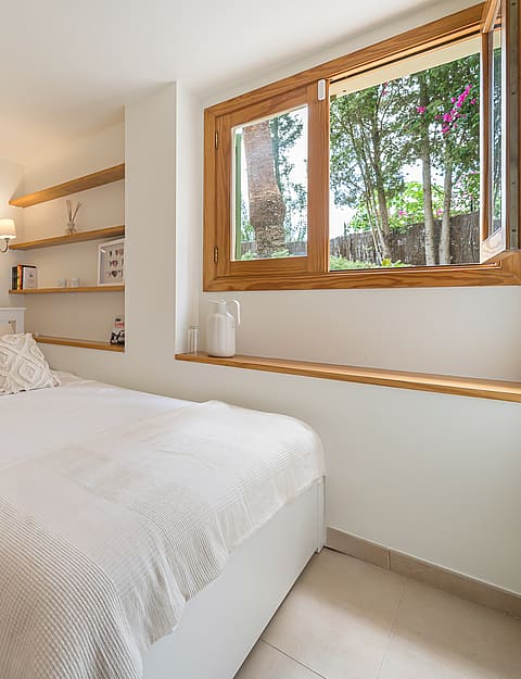 A bright bedroom featuring white linens, built-in wooden shelving, and a large timber-framed window facing garden trees.