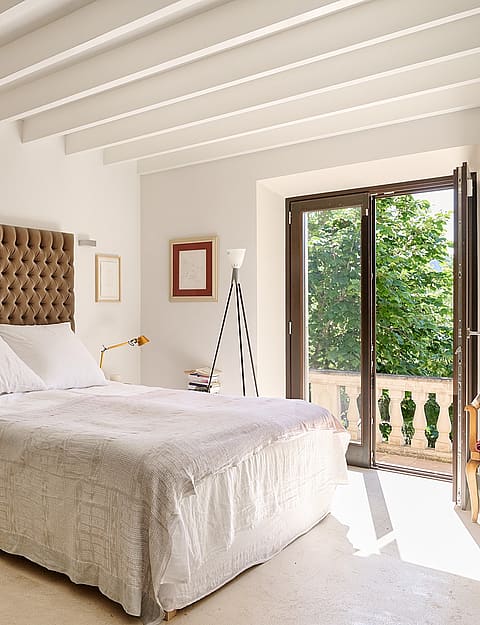 A bedroom with white walls, exposed wooden ceiling beams, a tufted brown headboard, and French doors leading to a terrace.
