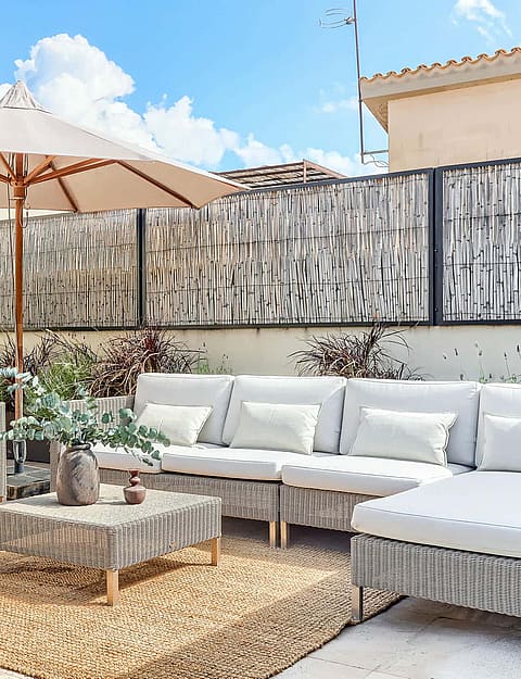 A rooftop terrace featuring a modular wicker sofa set with light cushions on a woven rug under a large beige umbrella.
