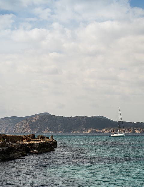 Rocky coastline meets turquoise Mediterranean waters with a white sailboat anchored before distant, arid hills.