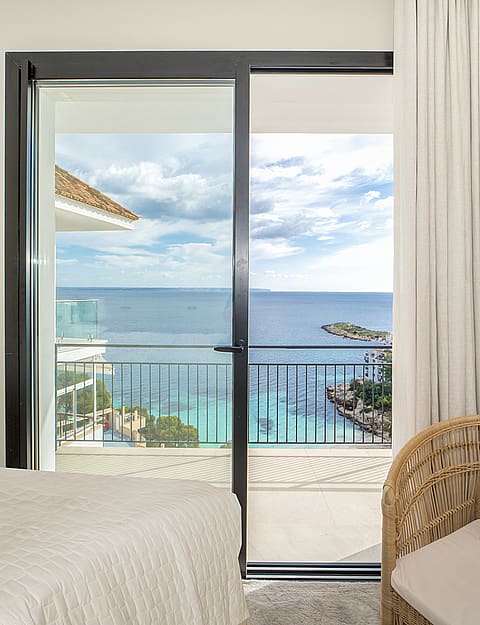 Interior view of a bedroom featuring sliding glass doors opening to a sea-view balcony with a woven chair in the foreground.