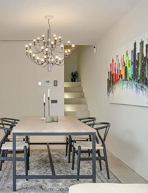 Interior view of a minimalist dining room with a stone table, black wooden chairs, a modern branching chandelier, and an abstract multi-colored painting on a white wall.