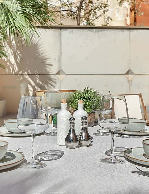 A dining table set for four on a stone terrace featuring cream-colored linens, ceramic bowls, and palm tree shadows.