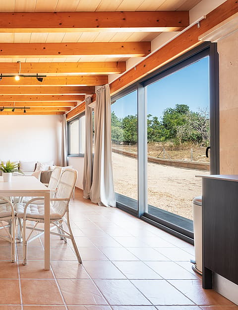 Comedor con mesa y sillas blancas, suelos de baldosas y puertas correderas hacia el exterior.