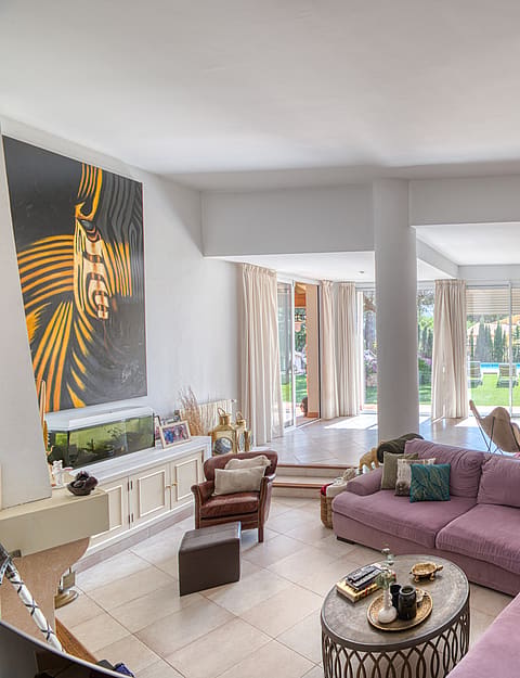 A spacious living room featuring light stone flooring, a large mauve sectional sofa, and access to a garden terrace.
