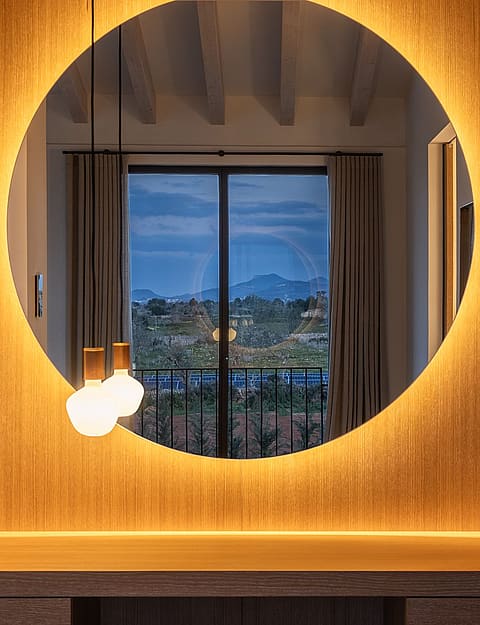 A backlit circular mirror over a wooden vanity table, reflecting a window with dusk mountain views in a Santanyí finca.