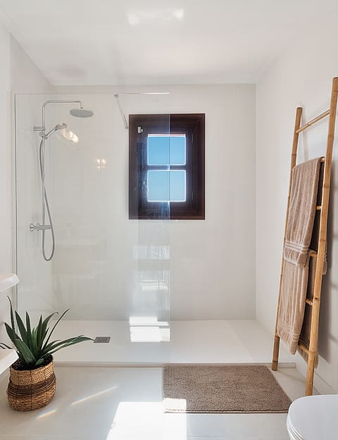 Baño blanco con ducha a ras de suelo, toallero de madera y detalles en fibras naturales.