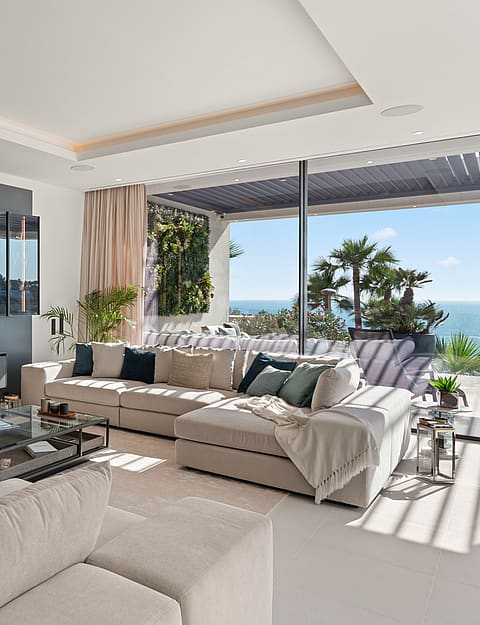 Modern living room with a charcoal grey feature wall, a wall-mounted flat-screen television, a beige sectional sofa, and floor-to-ceiling glass sliding doors opening to a terrace with sea views.