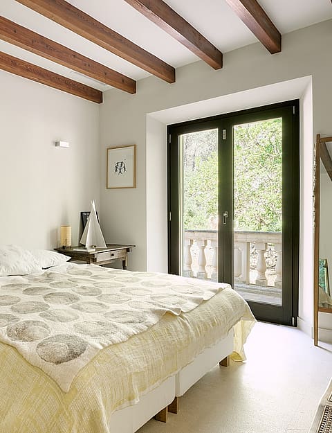 A bright bedroom with a queen bed, exposed dark wood ceiling beams, cream walls, stone flooring, and glass doors to a balcony.