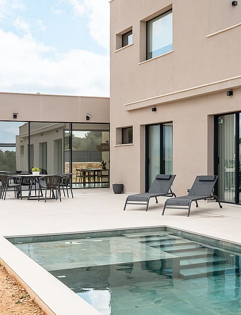 Rear facade of a modern beige villa showing a stone terrace, swimming pool with steps, and outdoor dining area.