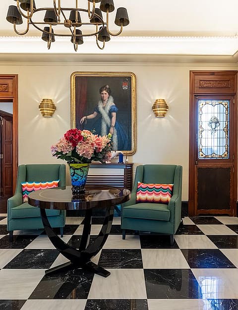 A formal entrance hall featuring black and white checkered marble floors, two teal armchairs, and a round central pedestal table.