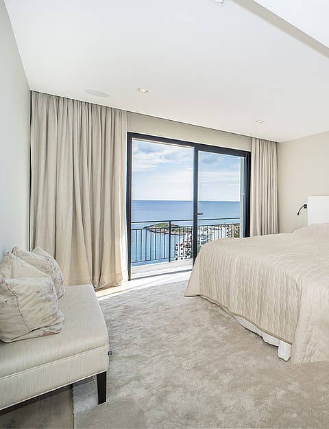 A minimalist bedroom featuring a large bed, neutral carpeting, light grey walls, and floor-to-ceiling glass sliding doors opening to a sea-view balcony.