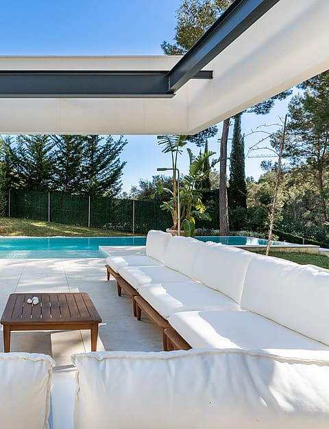 White cushioned outdoor seating under a steel pergola roof beside the swimming pool and pine garden