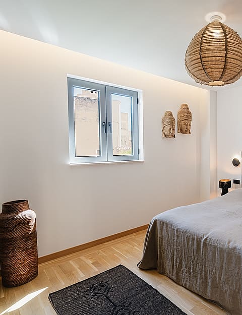 A minimalist bedroom featuring a grey upholstered headboard, light oak wood flooring, and a large woven pendant light.
