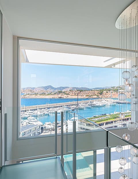 Top of a glass staircase featuring a cluster pendant light, with a wide window framing a view of Port Adriano harbor.