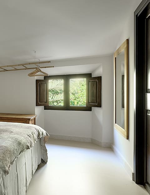 A bright bedroom in a finca with white walls, a bed featuring a patterned spread, a wooden chest, and an open casement window.