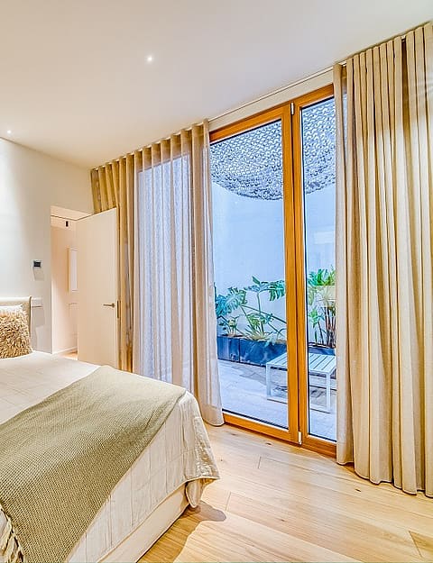 A modern bedroom with light wood floors, a neutral bed, and floor-to-ceiling glass doors opening to a private patio.