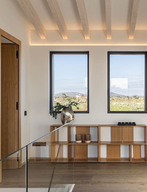 A bright hallway featuring wooden flooring, a low timber bookshelf, white walls with an abstract painting, and two windows showing mountain scenery.