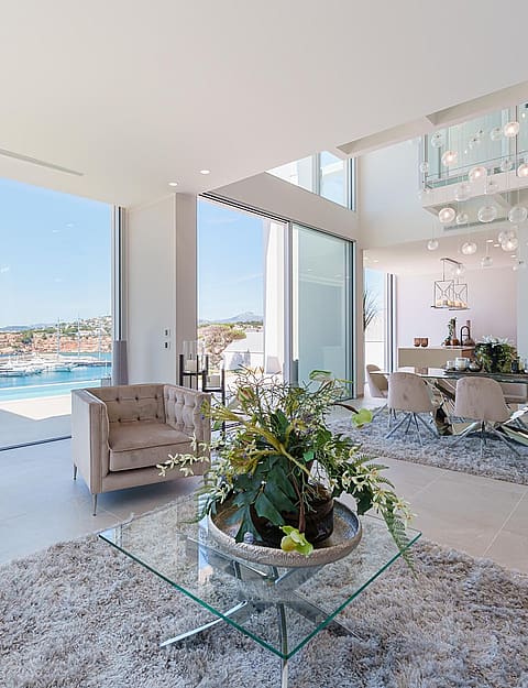 Modern open-plan living room with floor-to-ceiling windows overlooking Port Adriano marina, featuring tufted furniture.