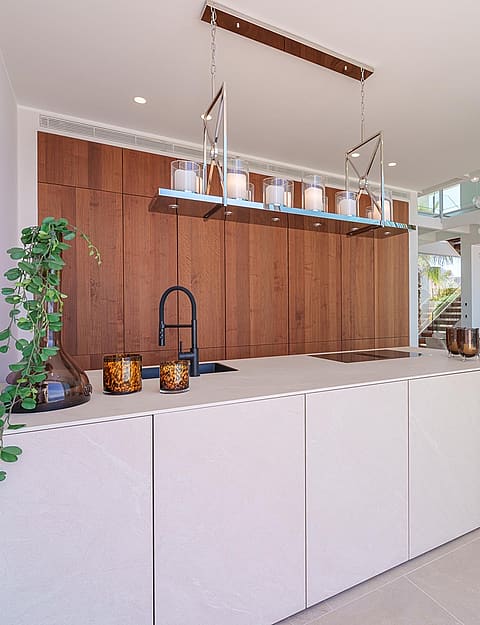 Modern kitchen island with matte white surfaces and dark wood wall-to-ceiling cabinetry, interior glass staircase visible in background.