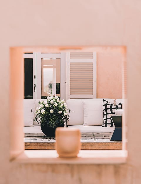 A square architectural opening in a pink stucco wall reveals a white sofa with patterned pillows and a potted plant on a terrace.
