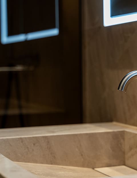 Close-up of a minimalist stone sink with a chrome wall-mounted faucet and backlit mirror in a modern bathroom.