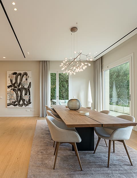 Dining room with a long wood table, modern sculptural chandelier, light oak flooring, and neutral walls in an open-plan villa.