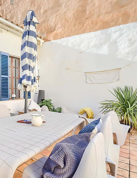 Outdoor terrace with a dining table and chairs, blue-striped sun umbrella, and blue shutters on a white building exterior.