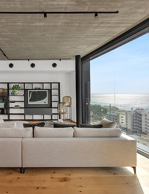 Wide angle interior view of an open-plan living room with light oak floors, raw concrete ceiling, and black track lighting.