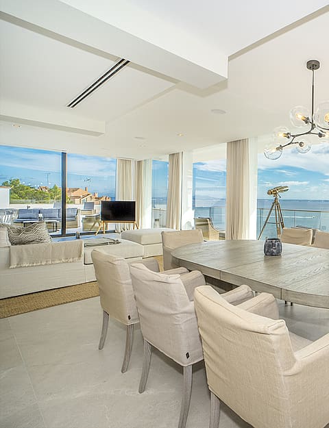 A dining area with an oval stone table and beige chairs, featuring a sculptural chandelier and a large glass-walled balcony.