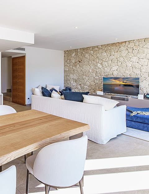 A bright open-plan space showing a kitchen with wooden cabinetry, a breakfast bar, and a living area featuring a stone wall.