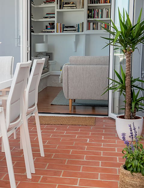A glass-topped dining table with white chairs on a terracotta-tiled balcony, overlooking mountain views from the apartment.