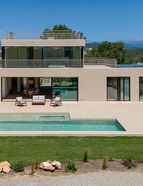 Exterior view of a modern two-story villa with a rectangular swimming pool, stone terrace, and distant mountain backdrop.