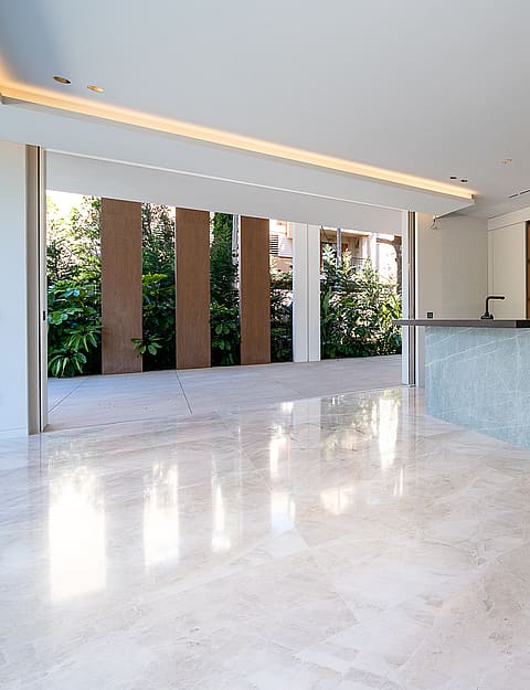 Modern kitchen featuring a large grey stone island, handleless white cabinets, and polished light marble floors. A wide glass door opens to a stone terrace and garden.