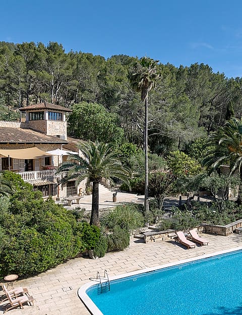 Wide angle view of a stone finca with terracotta roof and a swimming pool in a lush, tree-filled garden in Alaro.