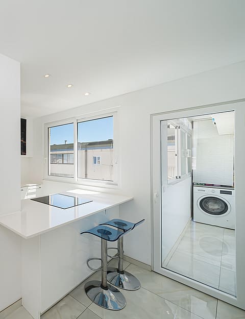 Modern white kitchen featuring a breakfast bar with two blue bar stools, tiled flooring, and an integrated oven.