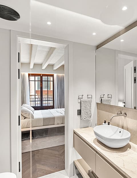 Modern bathroom featuring a stone vanity and white vessel sink, looking into a bedroom with exposed wooden beams.