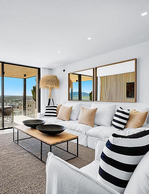 Modern open-plan living room with white sofas, wooden wall paneling, and large glass doors opening onto a terrace with mountain views.