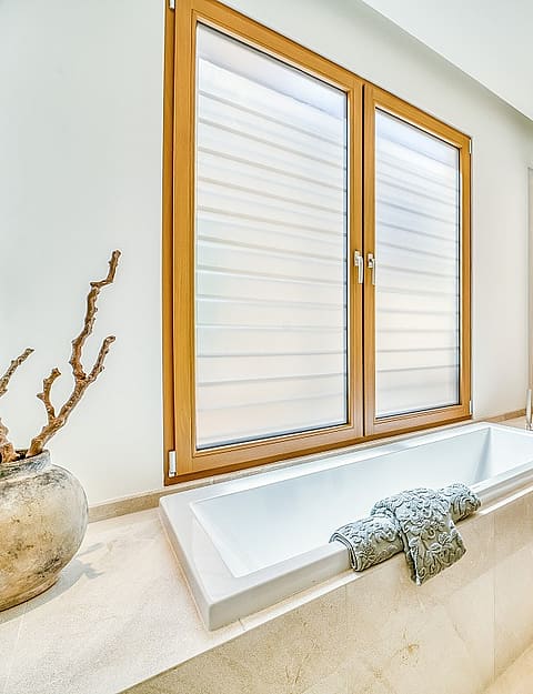 Modern bathroom featuring a rectangular white bathtub, wooden vanity, and large windows with blinds overlooking the city.