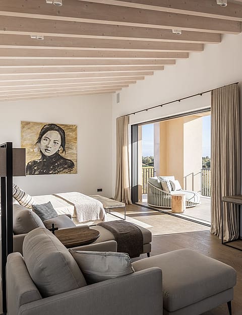 Spacious bedroom with white walls, wooden beam ceiling, gray modular seating, and glass doors opening to a sunlit terrace.
