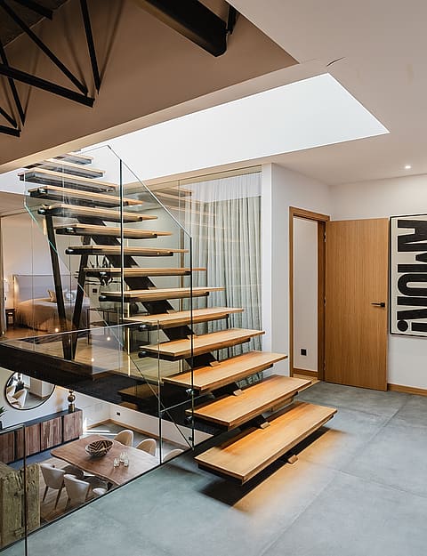 A modern floating staircase with wood treads and a glass railing connects levels in an apartment, viewed from a landing.