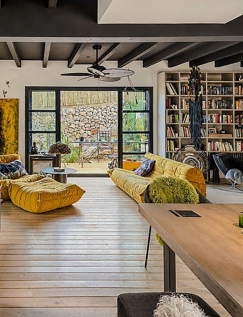 A living area with light wood floors, a large yellow sofa, and a wall-to-wall integrated bookshelf opening onto a stone terrace.